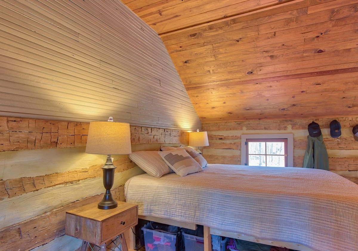 The upstairs bedroom under a sloping white-painted board ceiling, with a bed, a lamp on a small table, log walls and a small window.