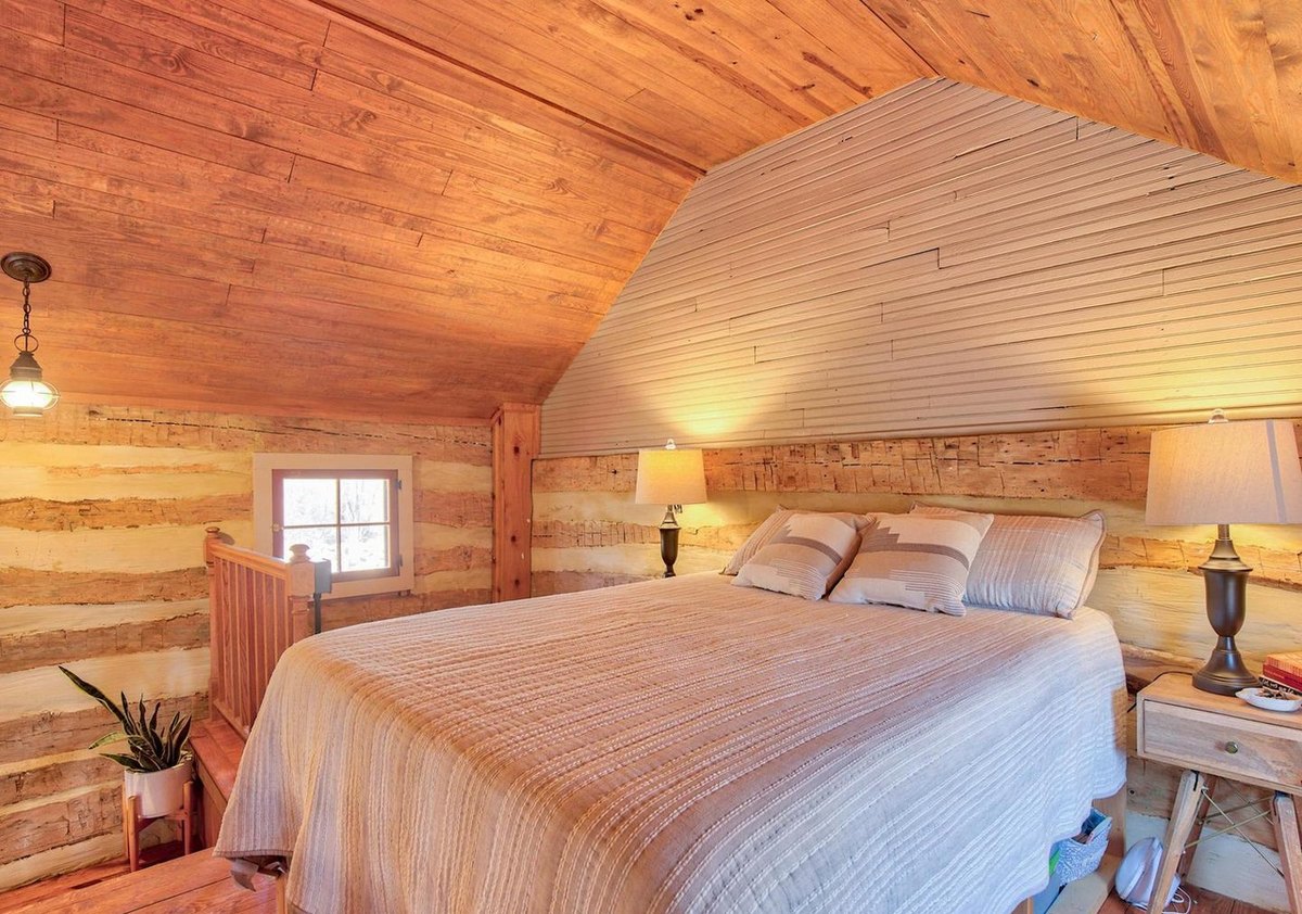 The upstairs bed seen head-on between two lamps on small tables, under a sloping ceiling, with a small window and the stair post at the left.