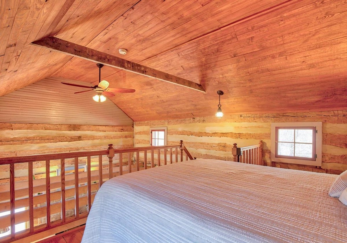 The upstairs room toward its gable end: a board ceiling with a ceiling fan and hanging light, small windows in the log walls, and a red stair railing beside the bed.