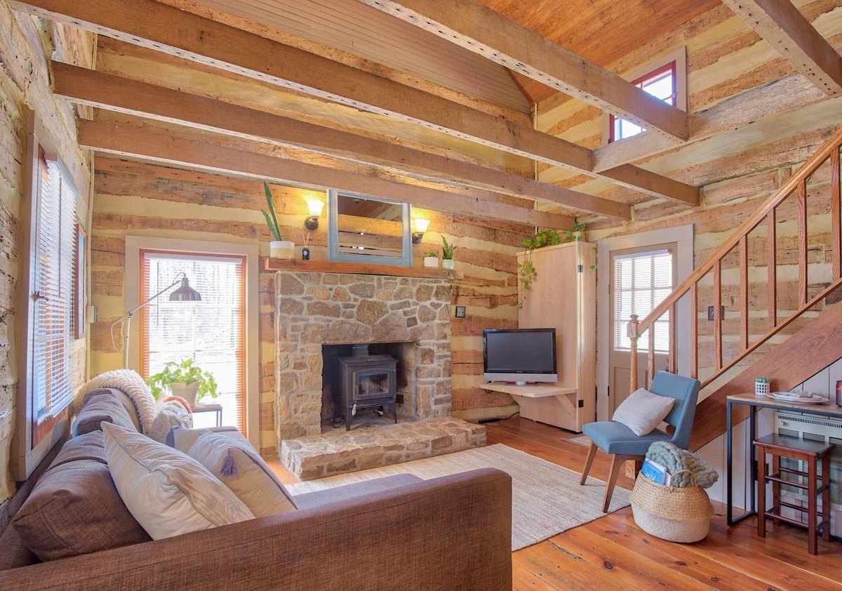 The main room with a sofa in the foreground, the stone fireplace and wood stove, a television, a blue chair and the stair along the log wall, under exposed beams.