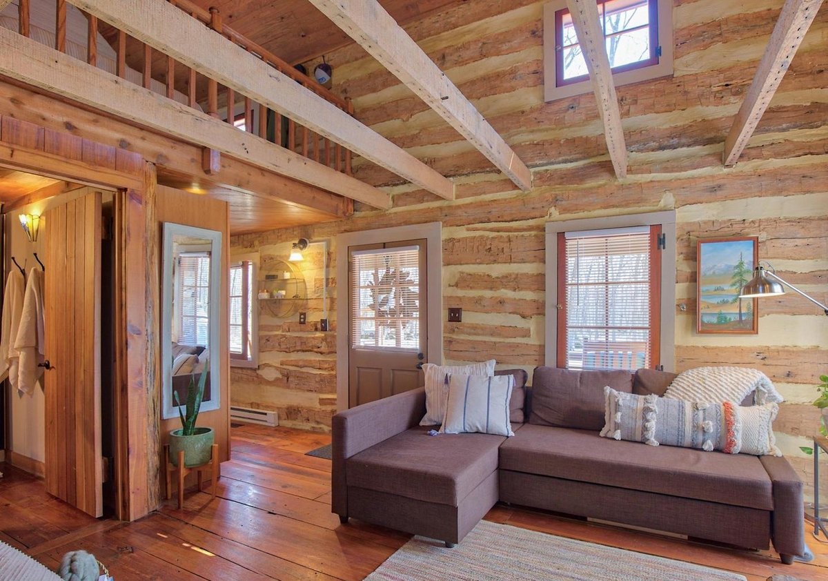 The main room toward the front of the cabin: a sectional sofa under a window and the front door, a board door and mirror at the left, and log walls under exposed beams.