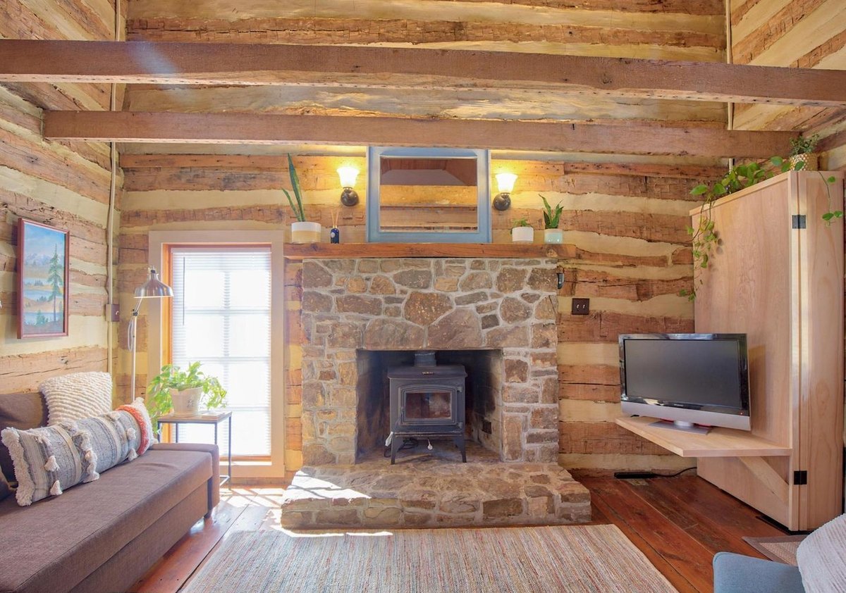 The fireplace wall of the main room: the stone fireplace with a wood stove on its hearth, a mirror and sconces on the mantel, a glazed door, and a wall-mounted television.