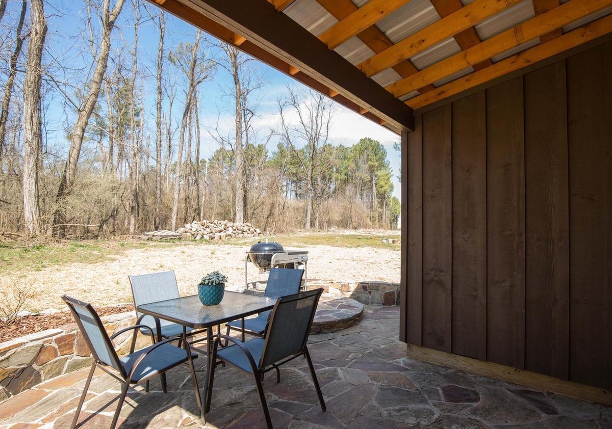 The covered back patio: a glass-topped table with four chairs on flagstones under the patio roof, a board wall at the right and the yard and woods beyond.