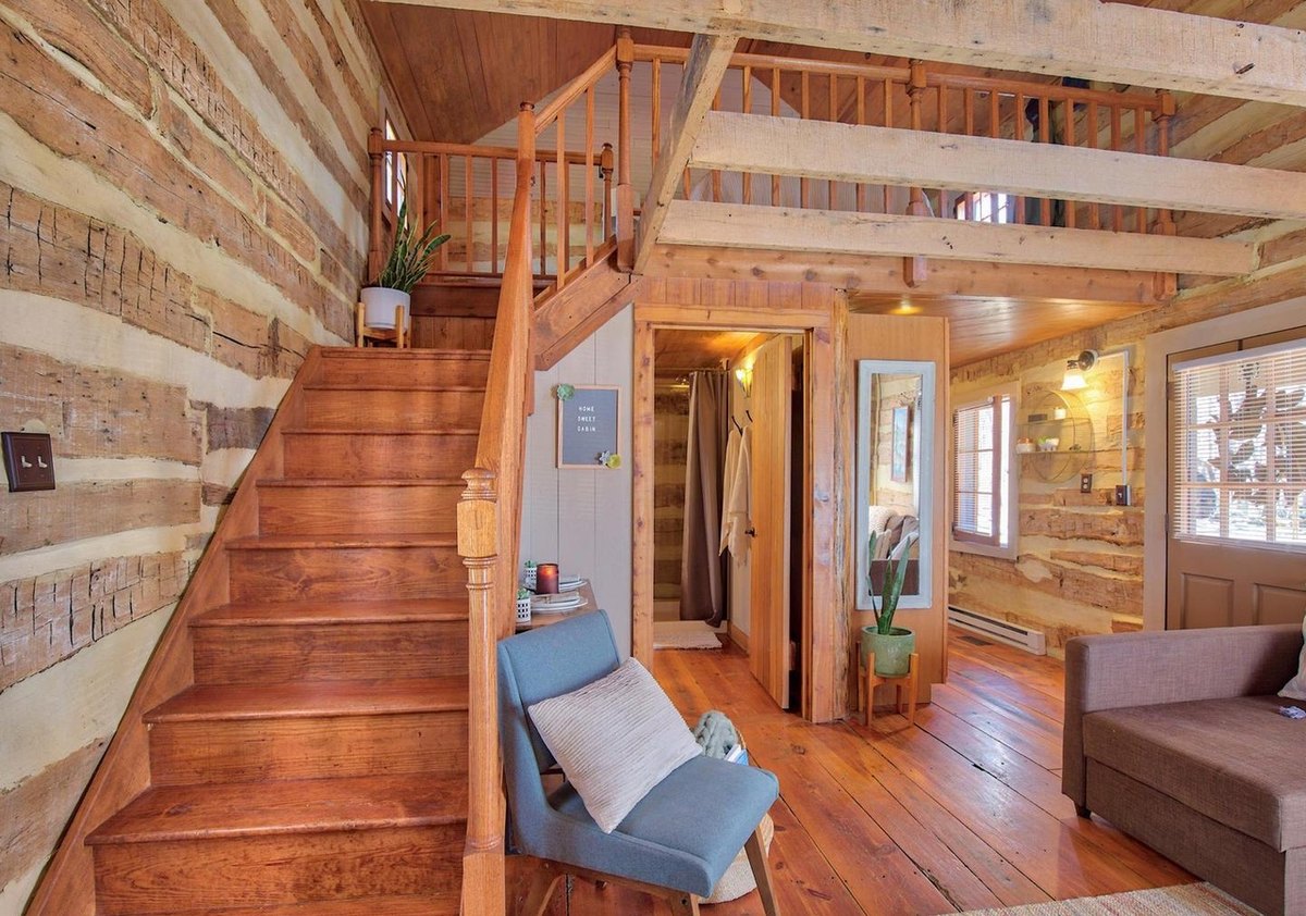 The wooden stair climbing to the loft along a log wall, with a blue chair, a doorway to the bathroom and the sofa at the right.