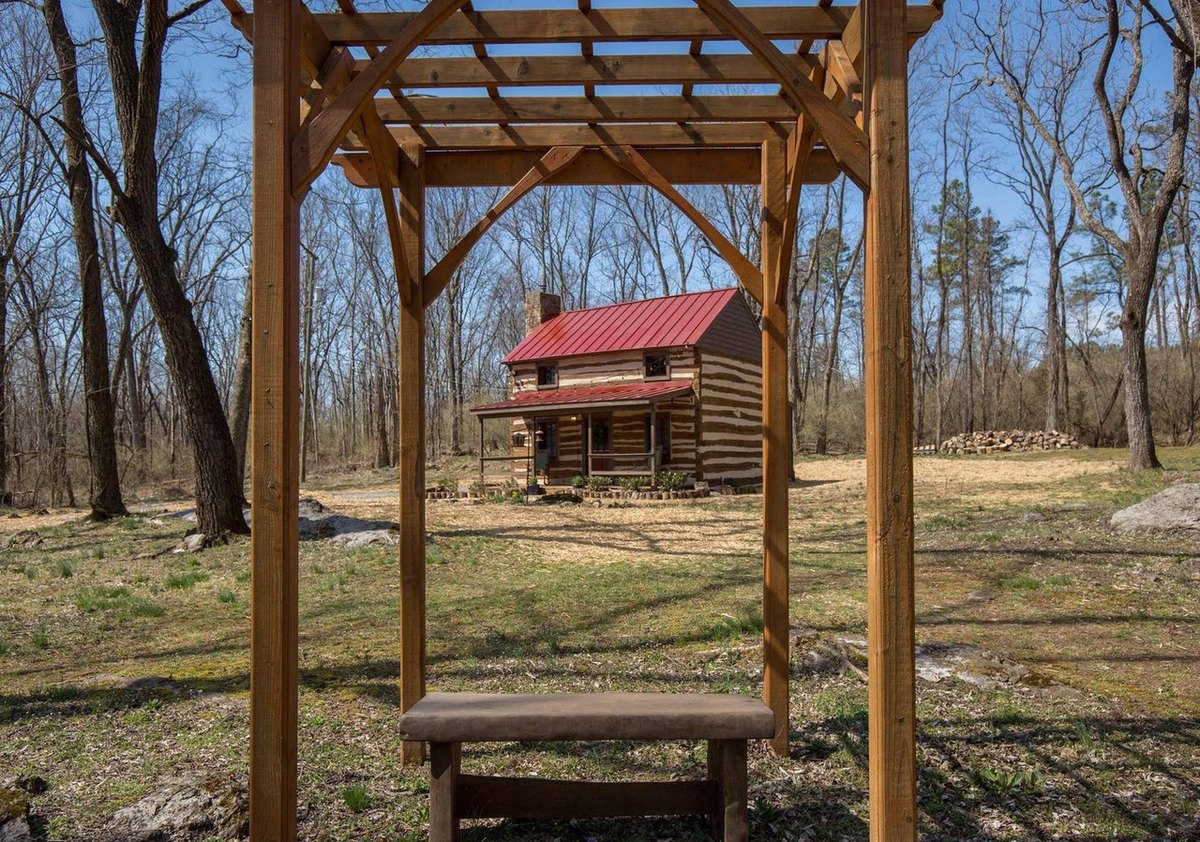 The cabin seen in the distance through a wooden arbor with a bench under it, across an open yard in early spring.