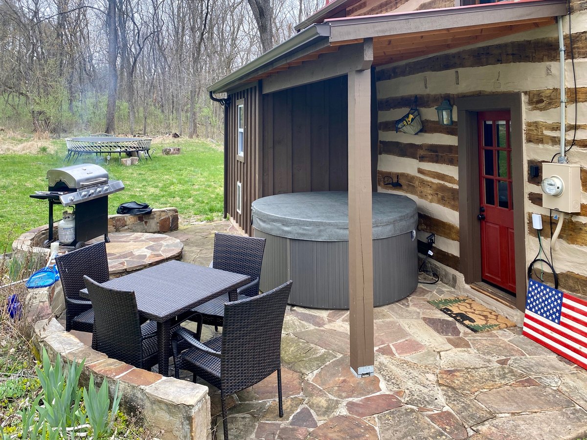The covered back patio beside the cabin: a hot tub, a wicker table and chairs, a gas grill and a painted American flag propped by the red back door, with the lawn and woods beyond.