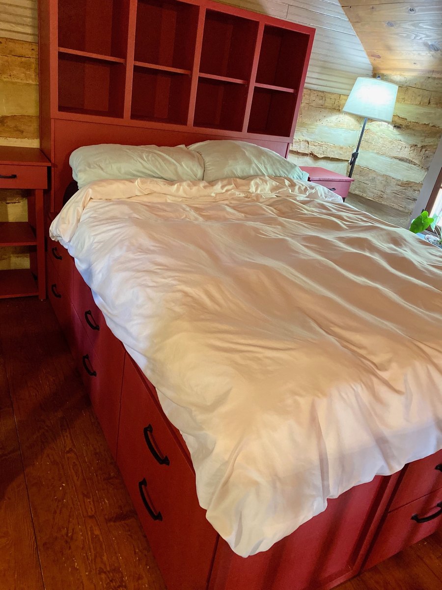 A bed with white linens and a red bookcase headboard with cubbies, in a room with log walls, a sloping ceiling and a lamp by the window.