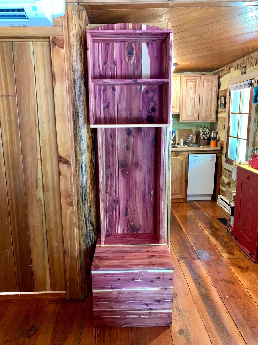 A tall new cedar cabinet with open shelves stands against a board wall, with the kitchen, a red cabinet and a log wall beyond, on a wide-plank floor.