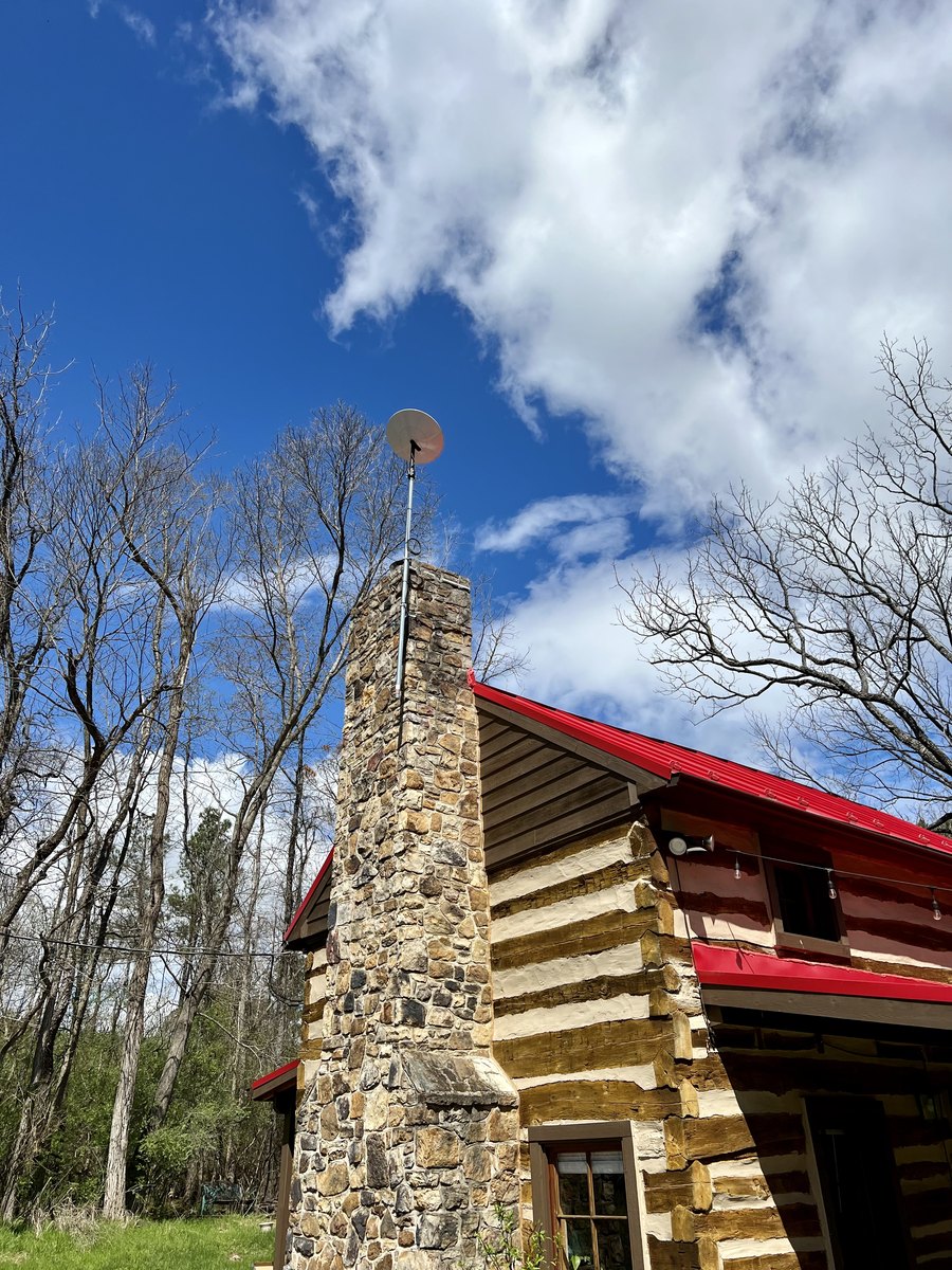 Looking up at the cabin's fieldstone chimney and red metal roof against a blue sky with white clouds, with hewn logs and a small upper window beside the chimney.