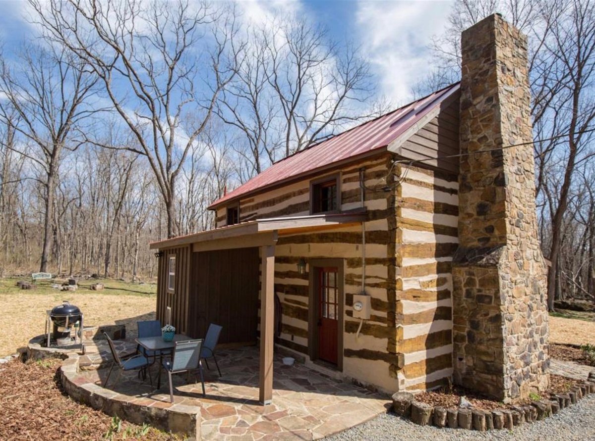The cabin from its back corner: a tall fieldstone chimney on the gable end, a lower shed-roofed addition with board siding, and a covered flagstone patio with a table, chairs and a kettle grill beside a red door.