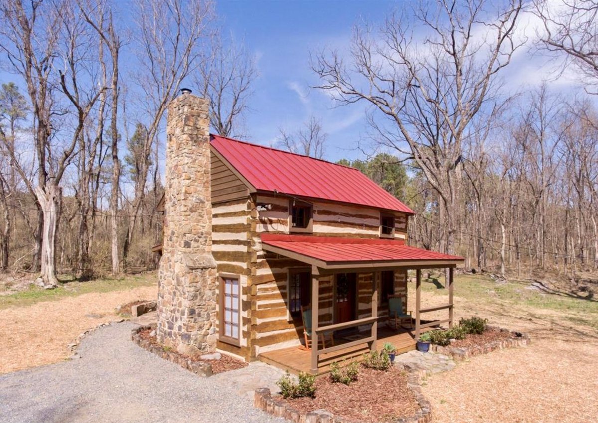 The cabin's chimney end: a tall fieldstone chimney beside a narrow glazed door, the red roof and the front porch turning the corner to the right, and a gravel path at its foot.