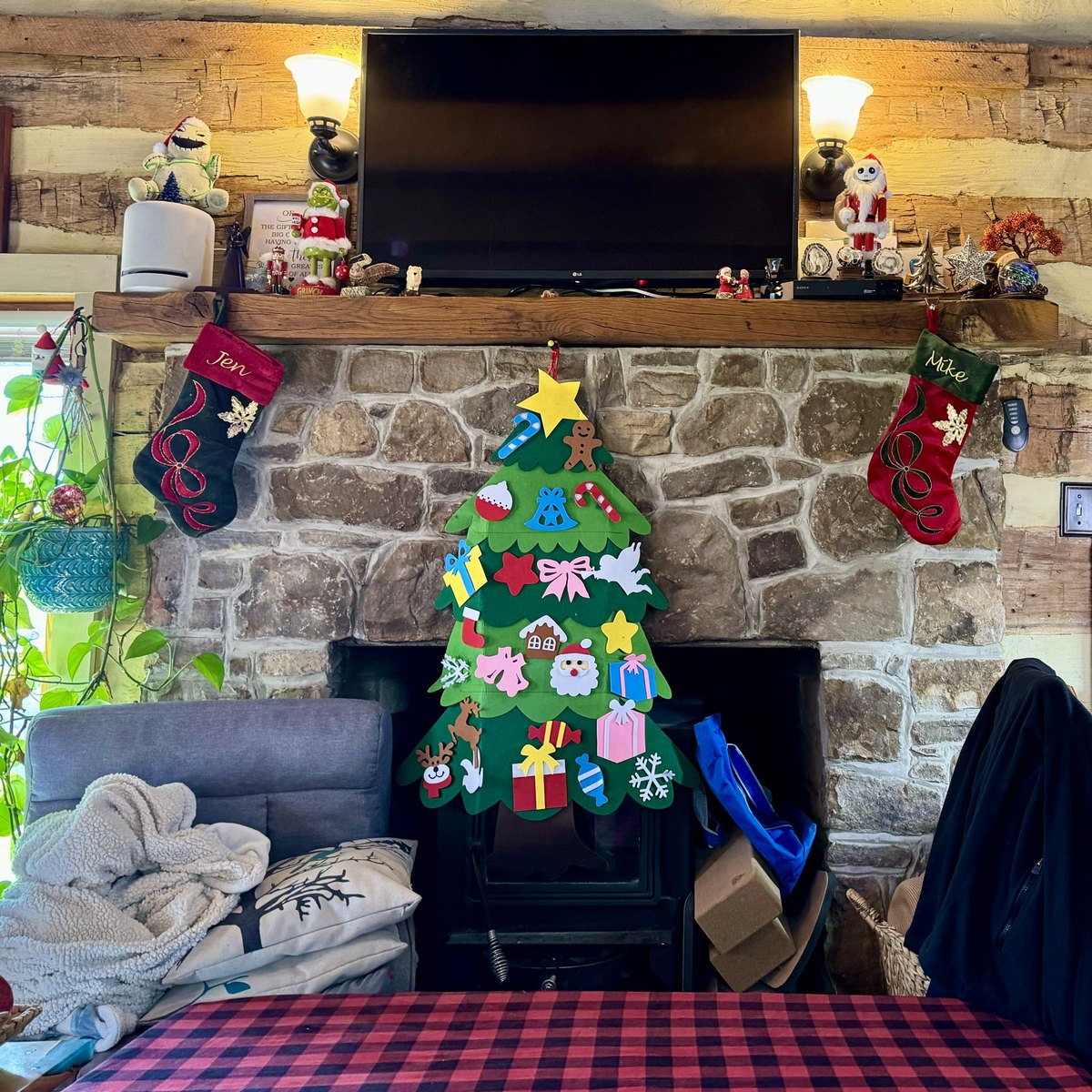 The stone fireplace decorated for Christmas: stockings hung from the wooden mantel, a felt Christmas tree over the wood stove, a television above, and a plaid tablecloth in the foreground.