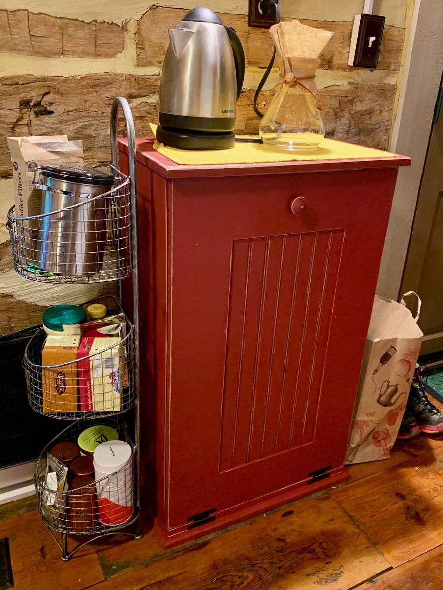 A closer view of the red cabinet with a kettle and a glass pour-over carafe on top, a wire basket stand of coffee and tea beside it, and the hewn logs of the wall behind.