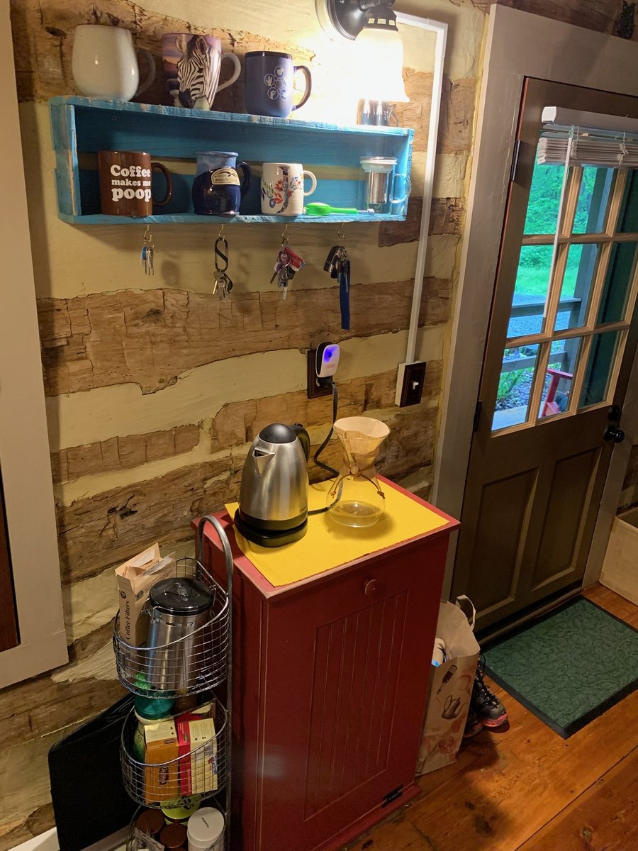 A corner against a log wall: a red cabinet with a kettle and a pour-over carafe on top, a blue shelf of mugs with keys hanging below it, and a half-glazed door to the porch.