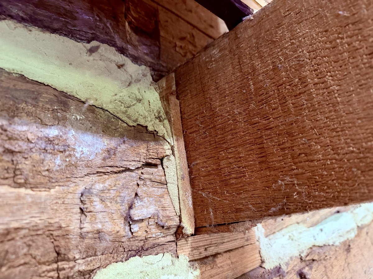 An angled view up into a corner where a beam with a finely textured sawn face meets the rough, weathered faces of the logs, with a thin board between them and pale infill along the log joints.