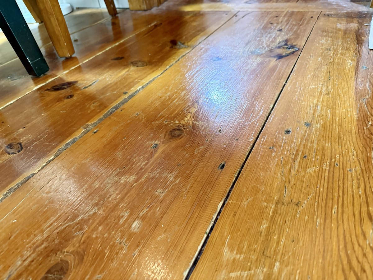 A low view along wide wooden floorboards under a glossy finish, showing knots, narrow gaps between the boards and scattered small dark marks, with the legs of a chair and table at the upper left.