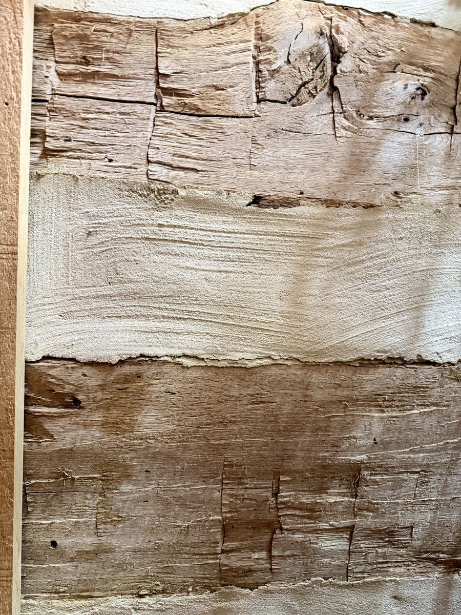 A straight-on view of an interior log wall: two log faces with rough, fibrous surfaces crossed by rows of short vertical cuts, and between them a wide band of pale infill with swirled trowel marks, beside the edge of a sawn board.