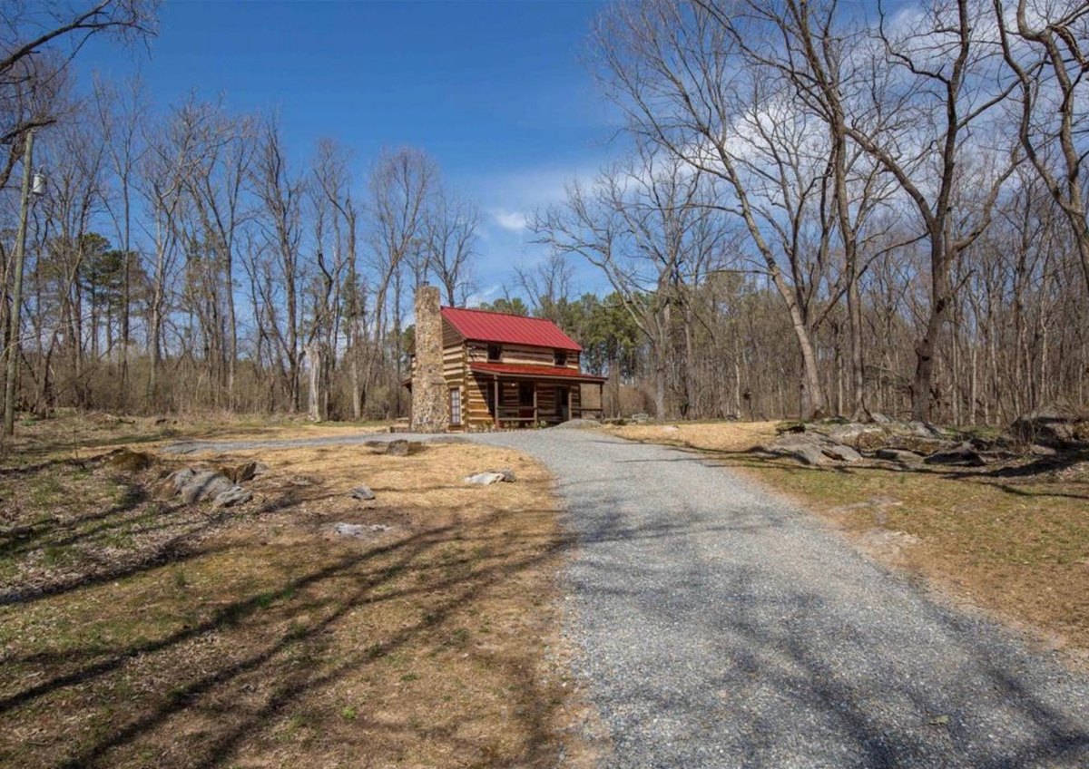 A gravel driveway curving through open ground toward the cabin, which stands at its far end among tall bare trees under a blue sky.