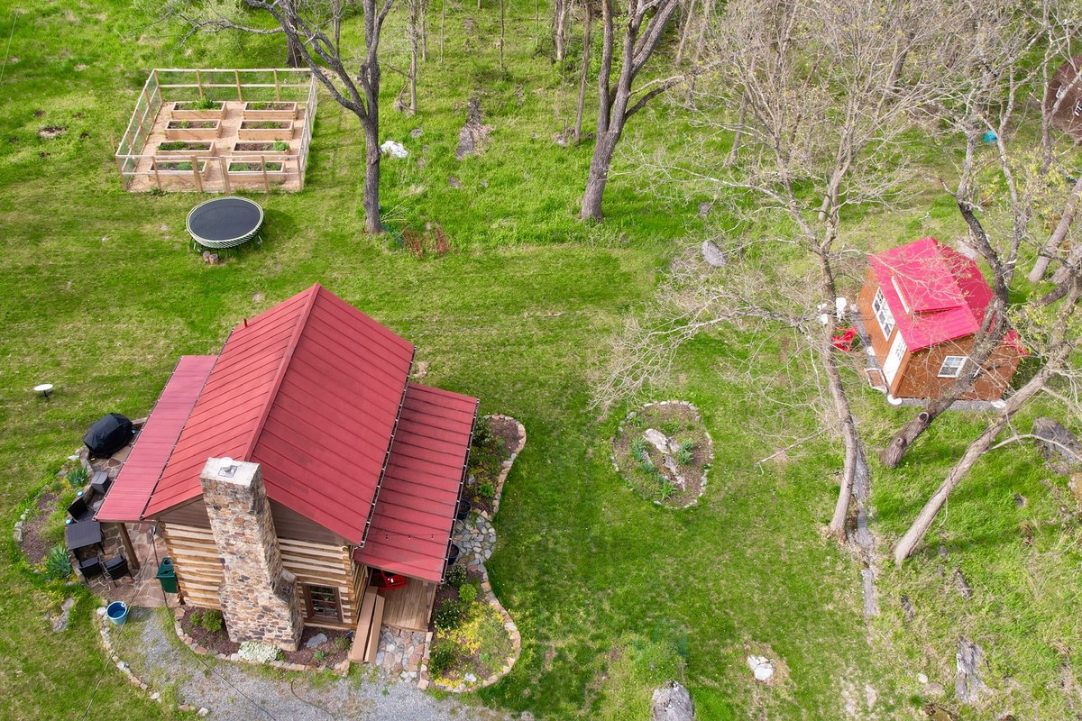 The cabin seen from above in spring: its red metal roof with lower roofs along both long sides and the stone chimney at one end, a patio and gravel path around it, and in the green yard beyond a fenced garden, a trampoline, a fire ring and a small red-roofed outbuilding among trees.