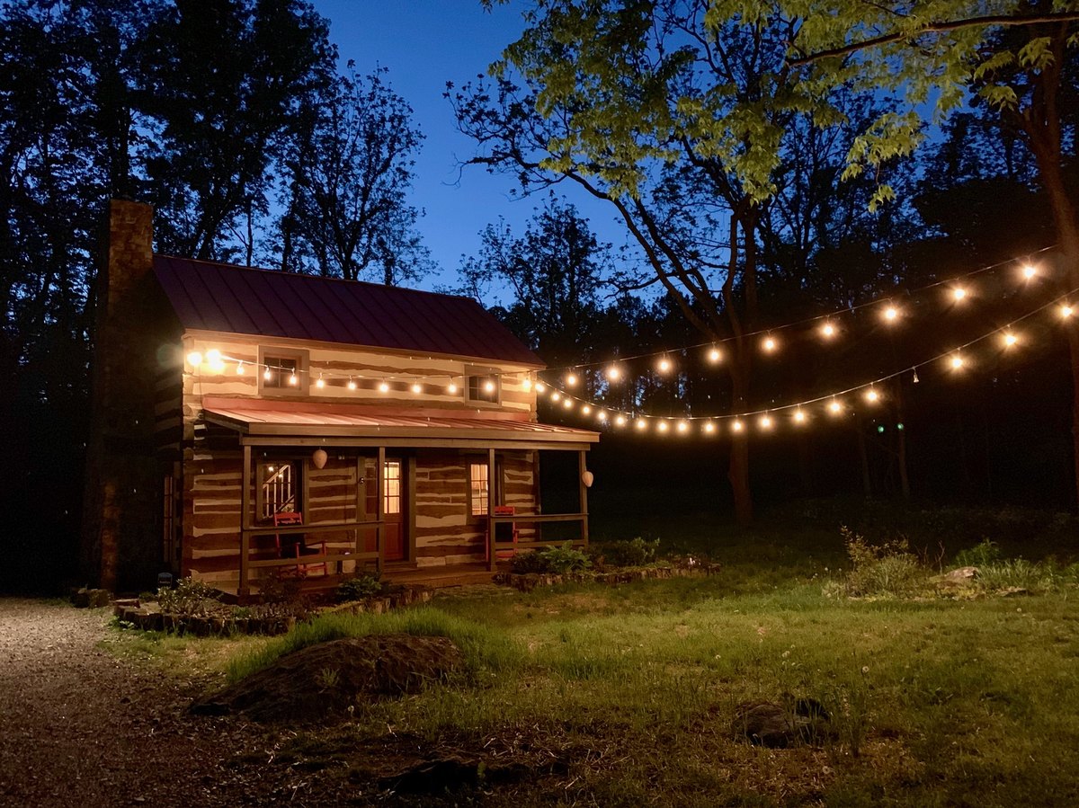 The cabin at dusk with its porch lit and strings of round bulbs running from the eaves out across the yard into the trees against a deep blue sky.
