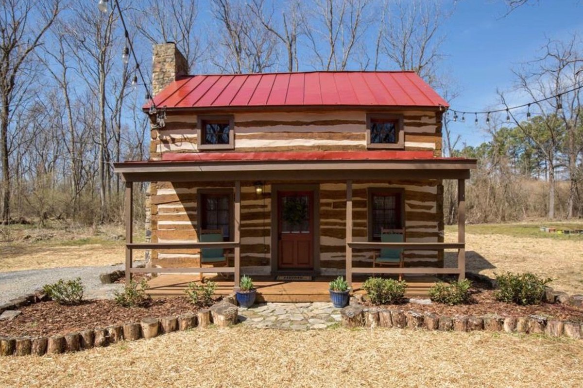 The log cabin seen straight on from the front on a sunny, leafless spring day: two stories of squared logs with pale chinking under a red metal roof, a covered porch across the front with two green chairs, a stone chimney at the left end, and new plantings edged with upright log sections.