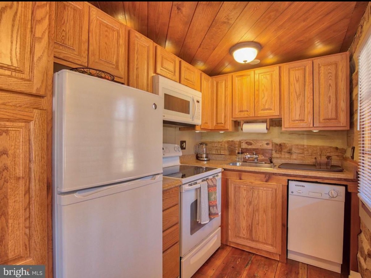 The small kitchen: oak cabinets, a white refrigerator, stove, microwave and dishwasher, a board ceiling, and a strip of log wall behind the counter.