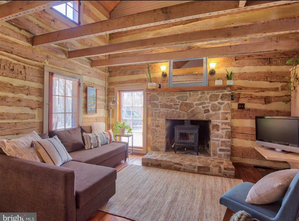 The main room: log walls with pale chinking, exposed ceiling beams, a stone fireplace holding a wood stove, a brown sectional sofa and a wall-mounted television, with a small window high in the gable.