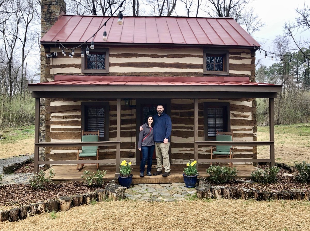 Michael Oak and Jennifer Riley standing together on the covered porch of the log cabin.