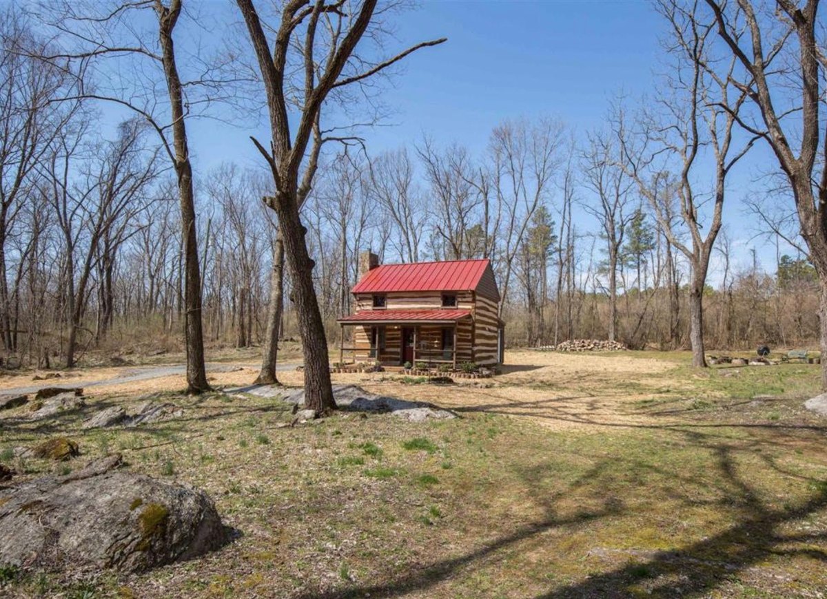 The cabin small in the distance across an open lawn scattered with rock outcrops, framed by tall bare trees under a clear blue sky.