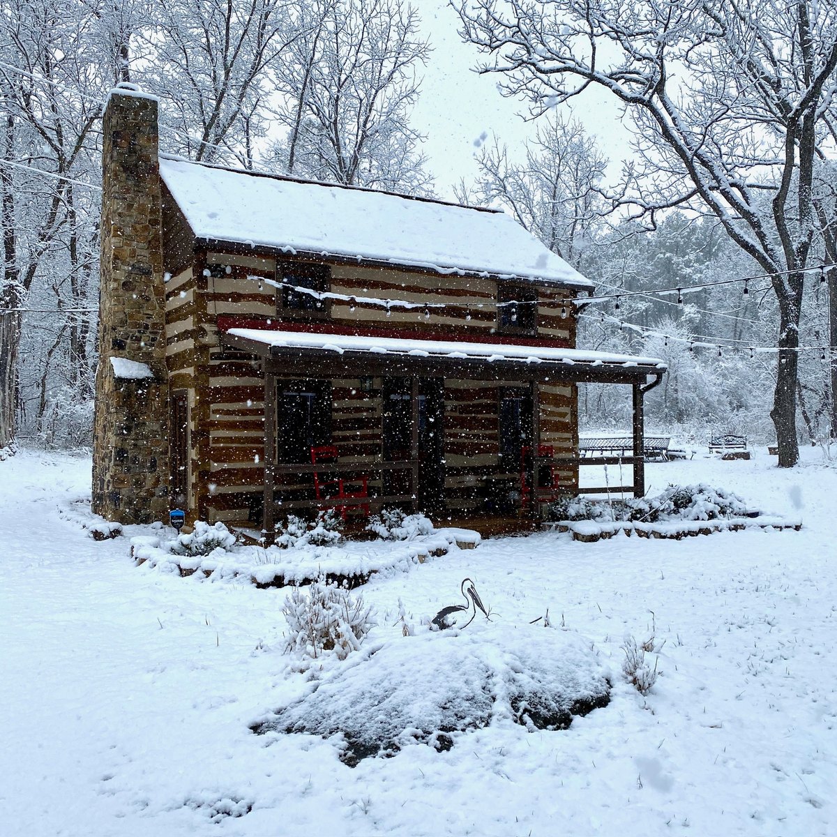 A two-story log cabin in falling snow, with pale chinking, a stone chimney and a covered front porch.