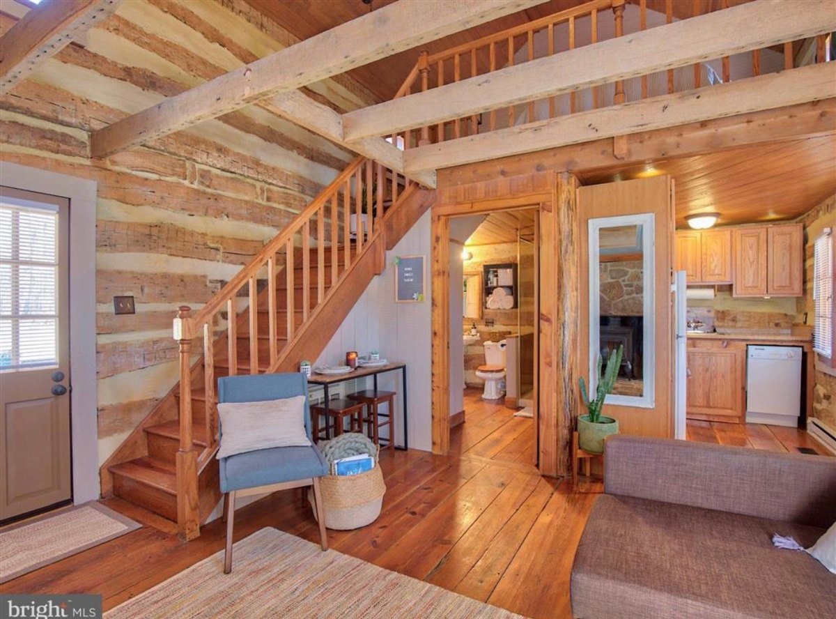 Inside the main room: an open wooden staircase rising along a log wall to a railed loft, a doorway to a small bathroom under it, and a view through to the kitchen, on wide pine floorboards.