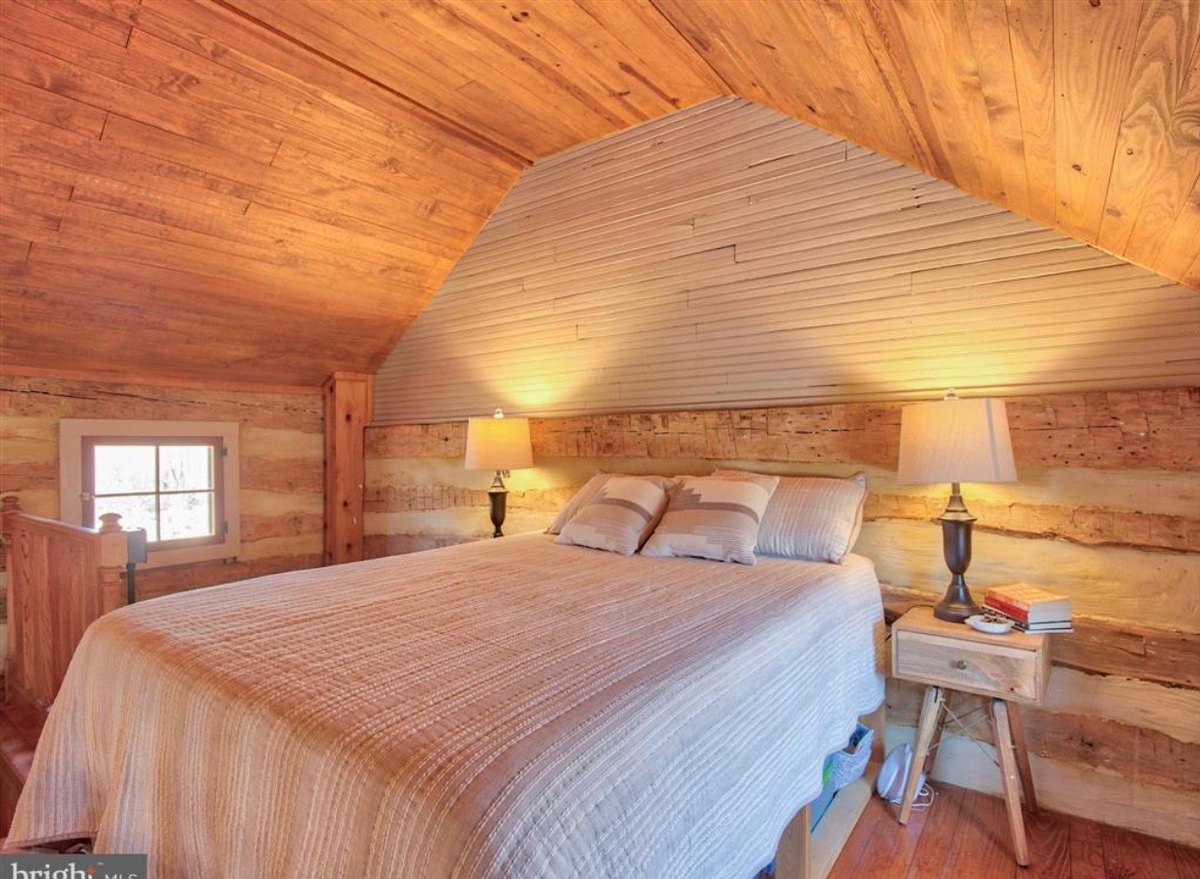 An upstairs bedroom under a sloping board ceiling, with a bed between two lamps against log walls and a small window beside the top of the stair.