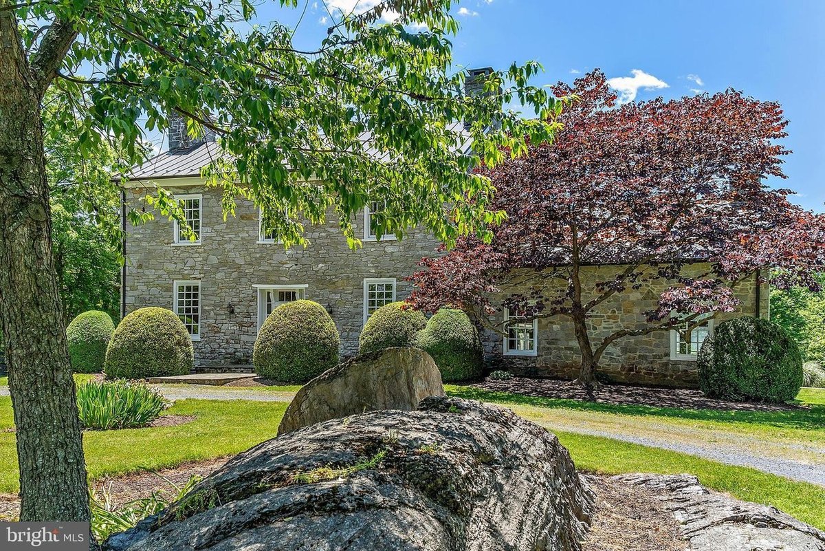 The same fieldstone farmhouse seen at an angle past a large lichen-covered rock, showing the two-story main block with its standing-seam metal roof and stone chimney and a lower stone wing to the right, half hidden behind a purple-leaved tree and rounded boxwood shrubs.